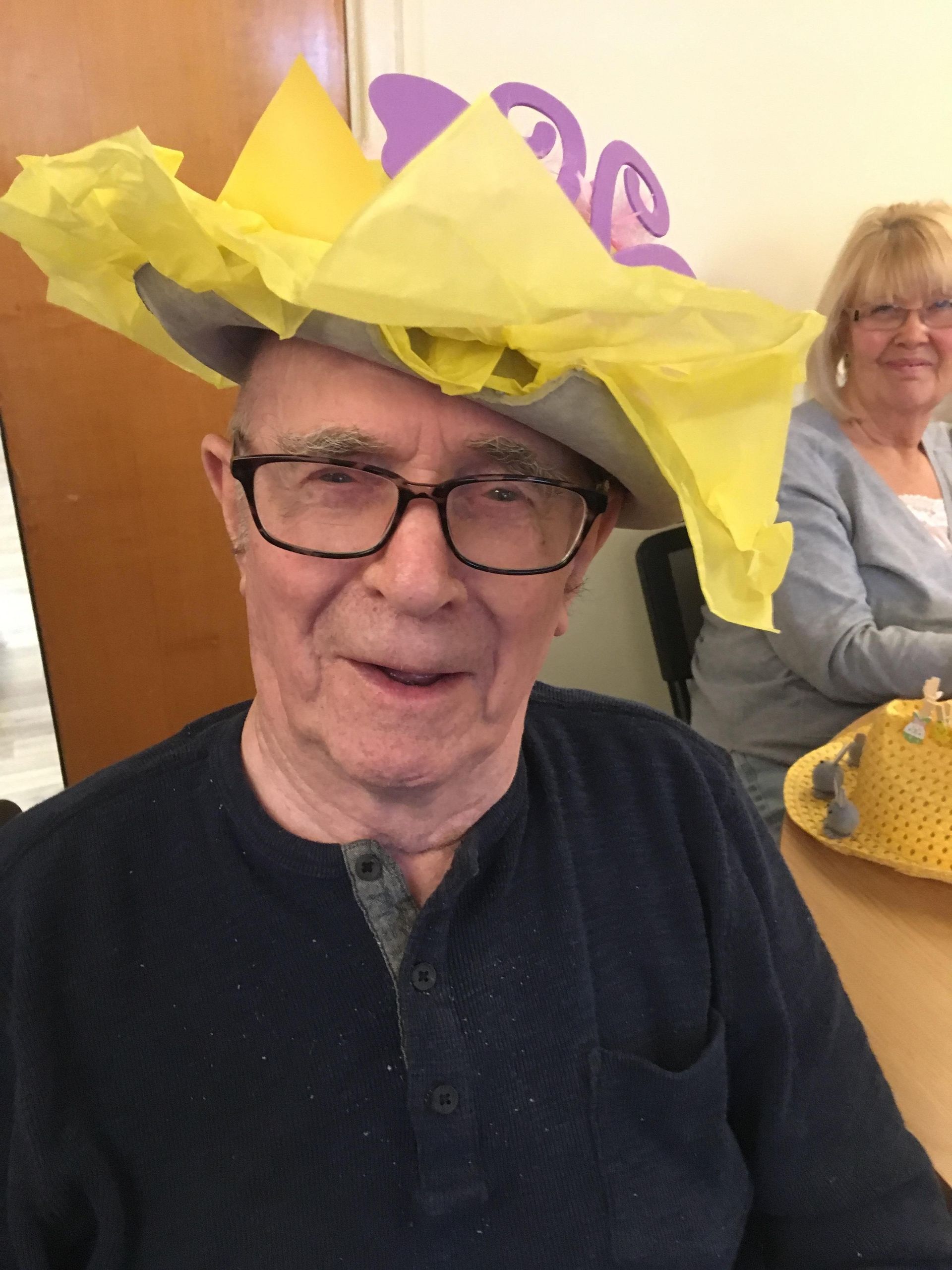 An older man wearing a whimsical yellow tissue paper hat, smiling, with a woman sitting in the background.